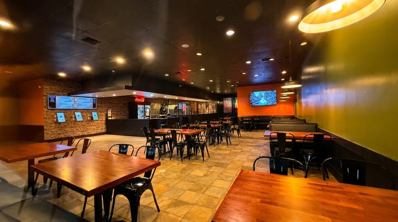 Empty restaurant interior with wooden tables, black chairs, and a counter with digital menus.