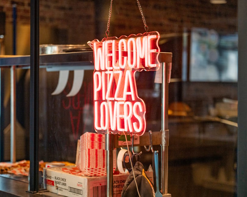 A bright red neon sign hangs, reading "WELCOME PIZZA LOVERS" above pizza supplies.
