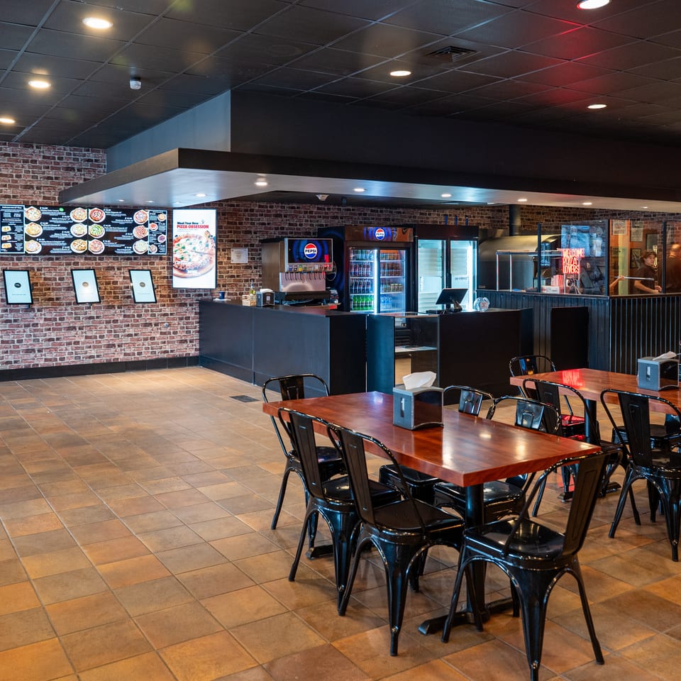 Interior of a restaurant with brick walls, black tables and chairs, and a counter with soda machines.