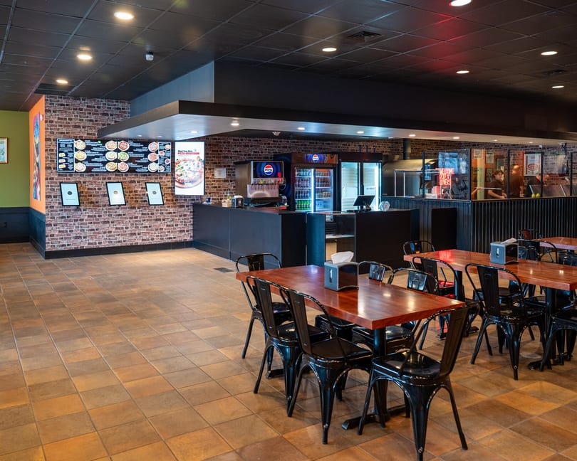 Interior of a restaurant with brick walls, black tables and chairs, and a counter with soda machines.