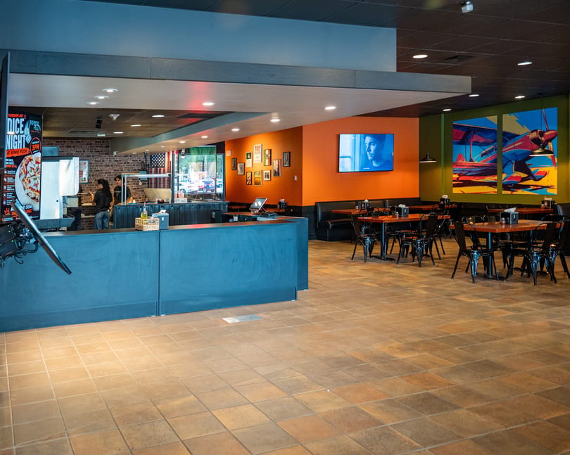 Spacious restaurant interior with orange and green walls, black tables, and a counter with two employees.