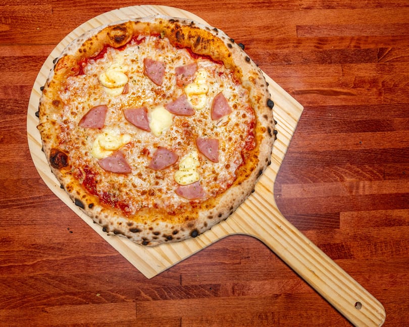 A whole pizza with ham, cheese, and a thick crust, on a wooden pizza peel.