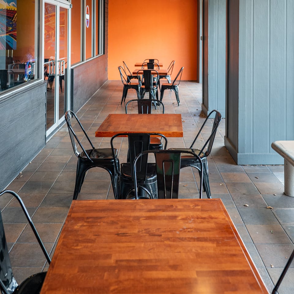 Outdoor patio with several wooden tables and black metal chairs, against an orange wall.