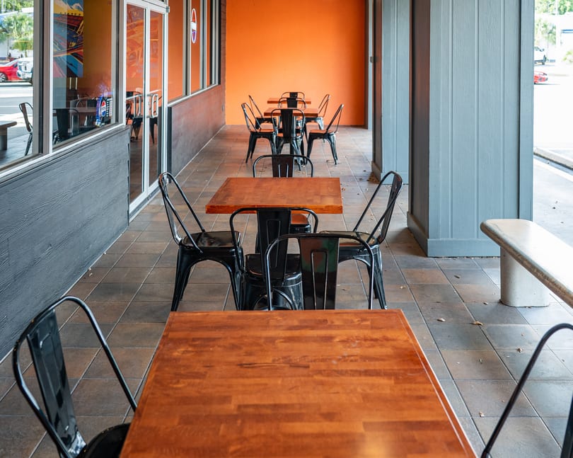 Outdoor patio with several wooden tables and black metal chairs, against an orange wall.