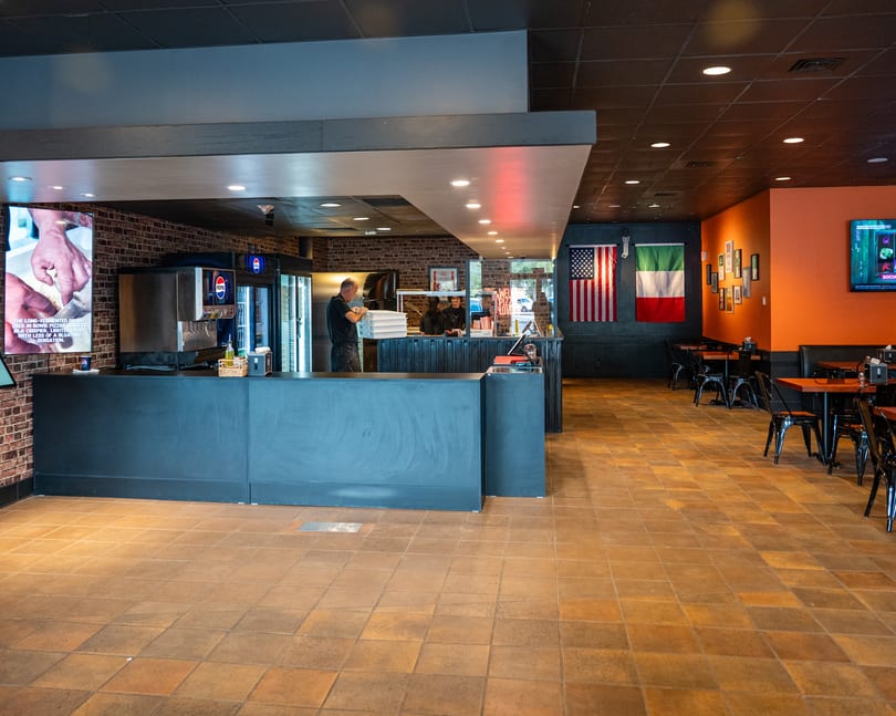 Interior of a pizza restaurant with a counter, kitchen area, and dining tables with flags on the wall.