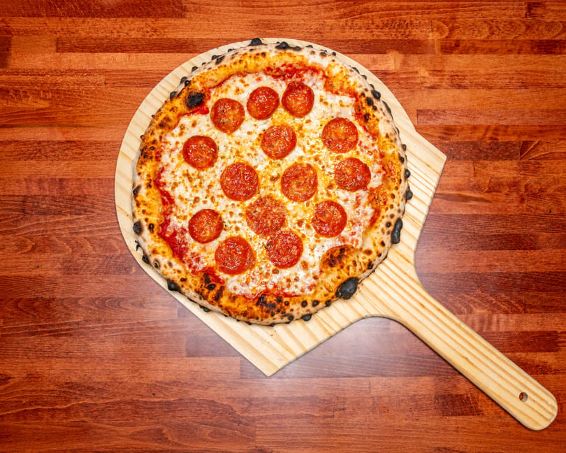 A whole pepperoni pizza with a charred crust on a wooden pizza peel, on a wooden table.