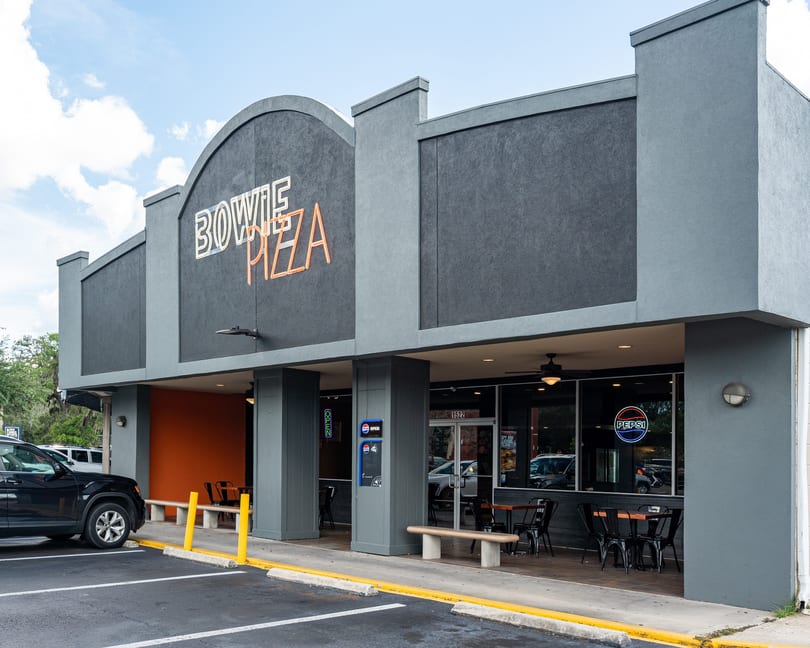 Exterior of Bowie Pizza restaurant with a dark gray facade, neon sign, and outdoor seating.