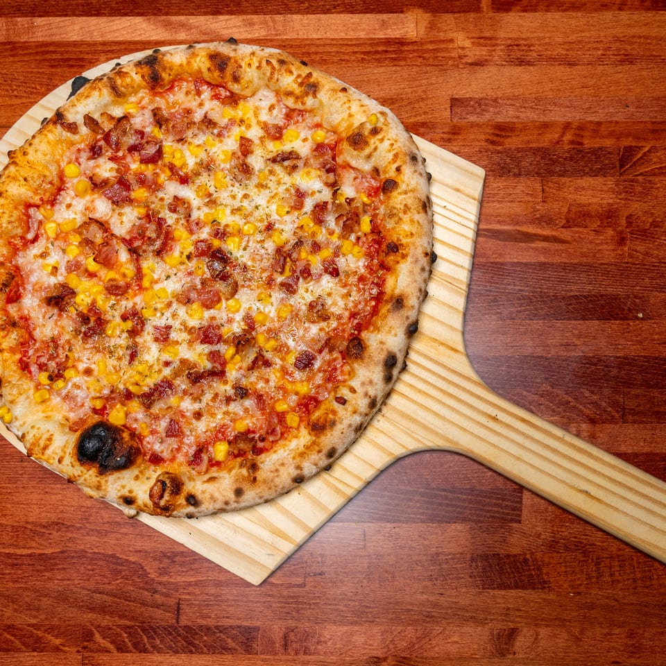A freshly baked pizza with bacon and corn toppings on a wooden pizza peel, on a dark wooden table.