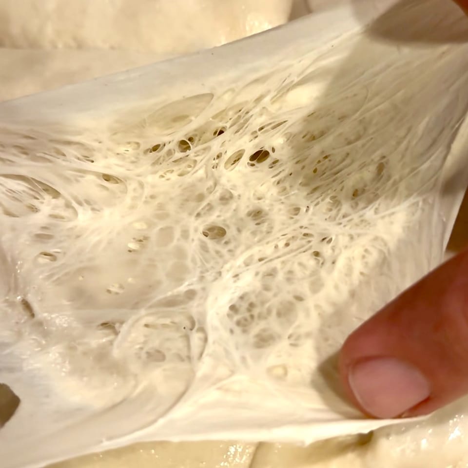 A hand stretches light-colored dough, revealing a web of gluten strands and air pockets.