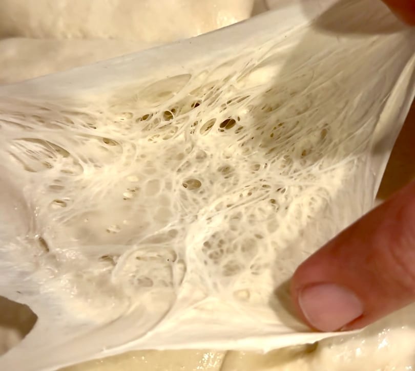 A hand stretches light-colored dough, revealing a web of gluten strands and air pockets.