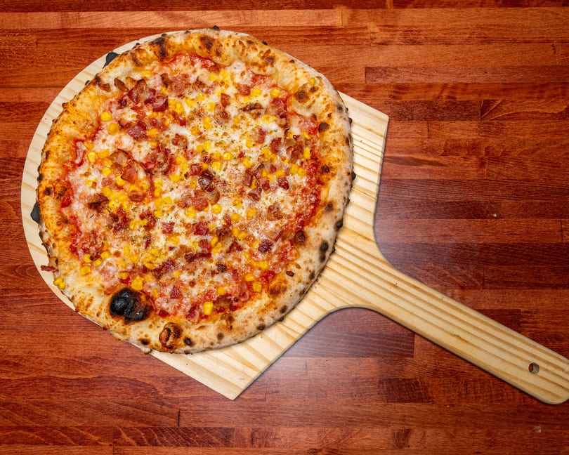 A freshly baked pizza with bacon and corn toppings on a wooden pizza peel, resting on a dark wood table.