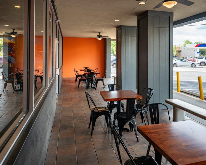 Outdoor patio with several empty tables and chairs, an orange wall, and a view of a parking lot.