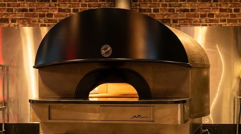 A large, black and bronze pizza oven with a glowing interior, set against a brick wall.