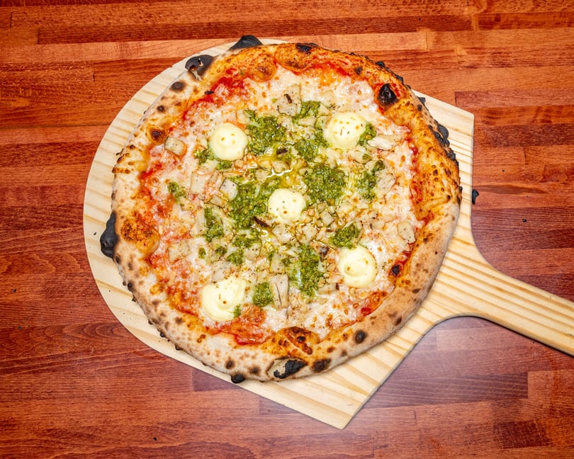 A round pizza with pesto, chicken, cheese, and dollops of white sauce on a wooden peel.