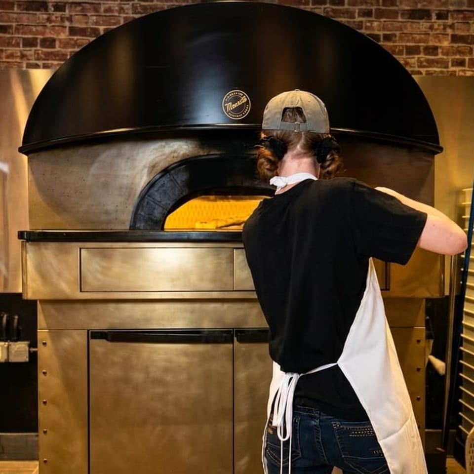 Chef in apron and hat placing a pizza into a large, glowing wood-fired oven in a brick kitchen.