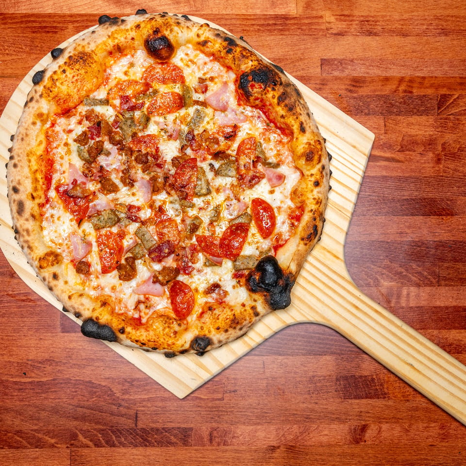 A freshly baked meat lover's pizza with a charred crust on a wooden peel, on a wooden table.