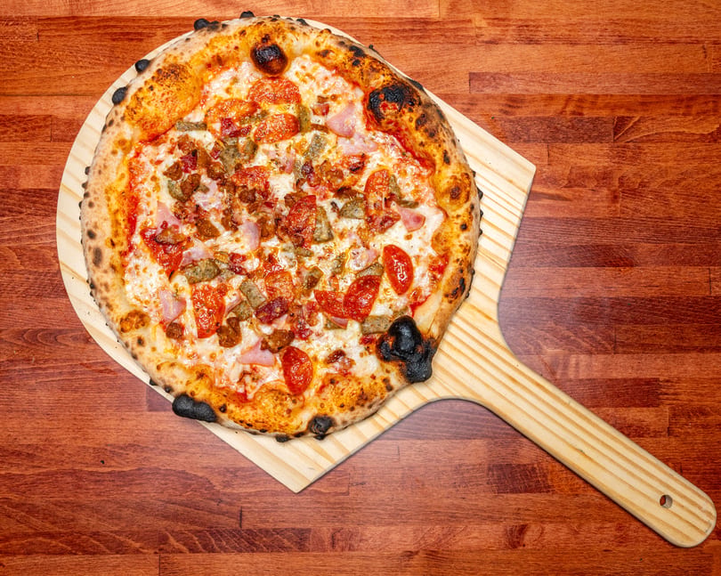 A freshly baked meat lover's pizza with a charred crust on a wooden peel, on a wooden table.